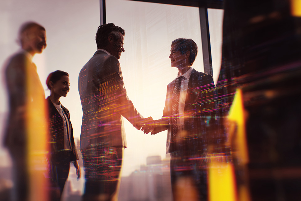 Two businesspeople shake hands in warm sunset light by a large window, with colleagues blurred in the background.